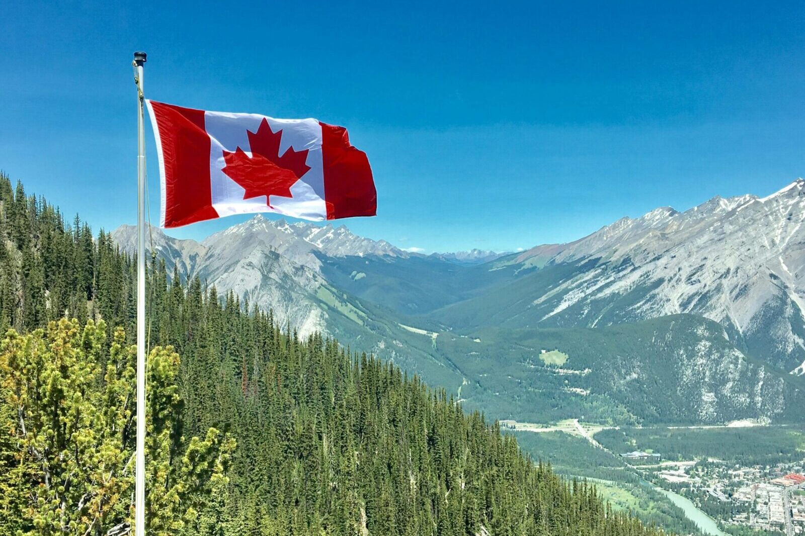Canadian flag waving atop a scenic mountain view with blue skies and lush greenery.